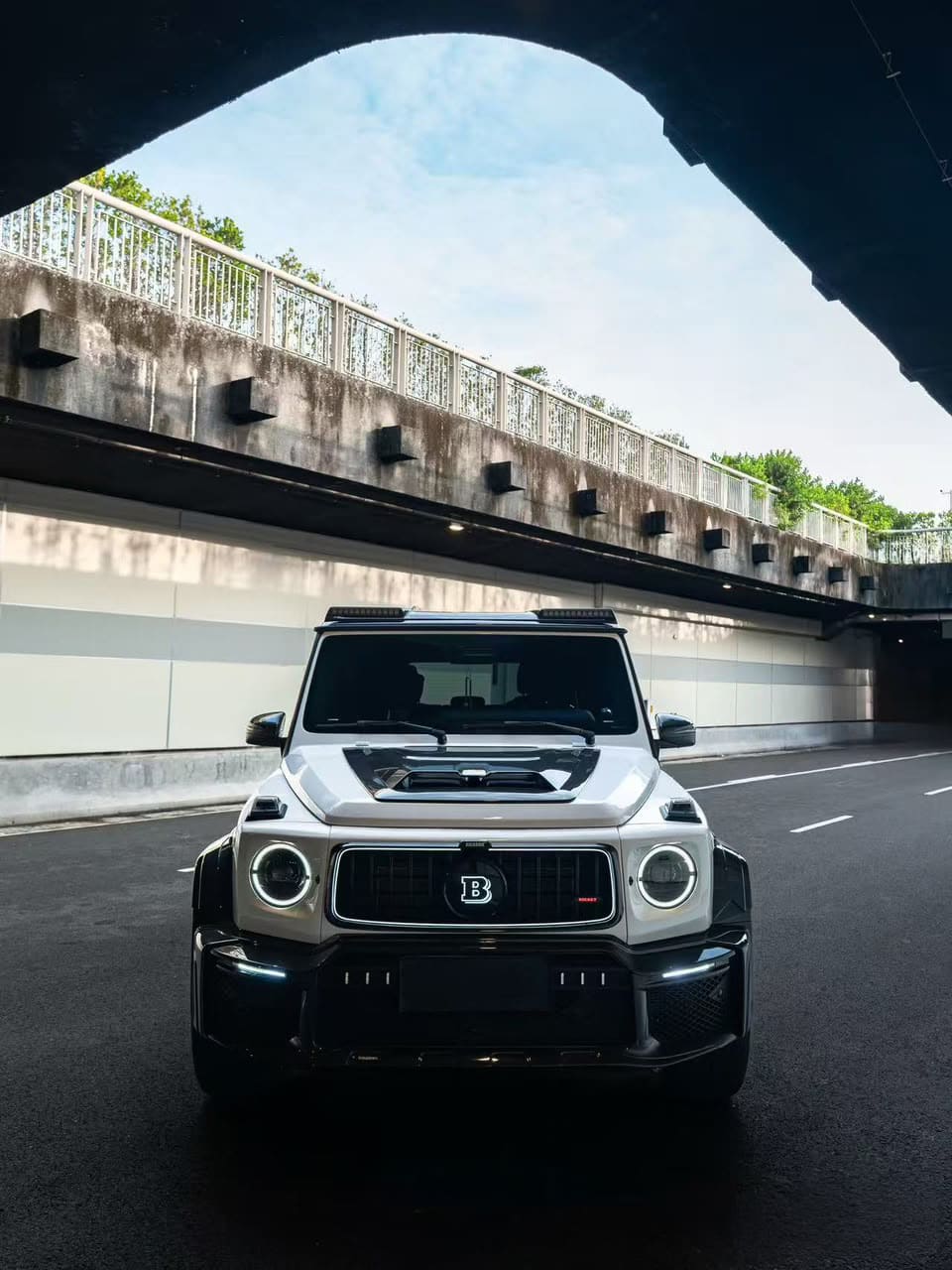 Straight-on front view of a white Mercedes-Benz G-Class W465 featuring a Brabus Rocket 900 style carbon fiber widebody kit, illuminated "B" emblem, and LED roof light bar.