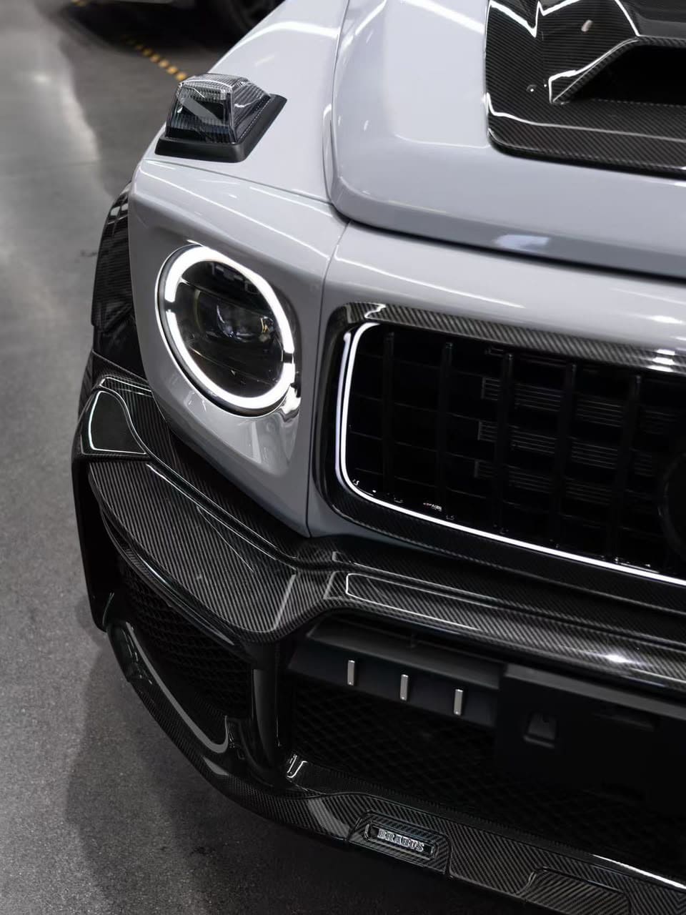 Close-up of Mercedes-Benz G-Class W465 front fascia showing gloss pre-preg carbon fiber bumper lip spoiler with Brabus badge and circular LED headlight.